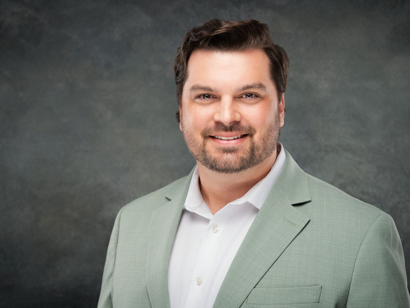 Smiling man with short dark hair and beard wearing a light green suit jacket and white shirt against a gray studio background.