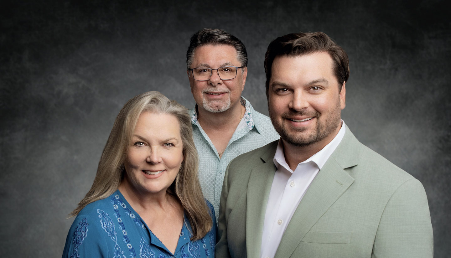 Portrait of a smiling woman with long blonde hair in a blue patterned blouse, a man with glasses and salt-and-pepper hair in a light blue shirt, and a man with short brown hair and beard wearing a light gray suit jacket and white shirt.