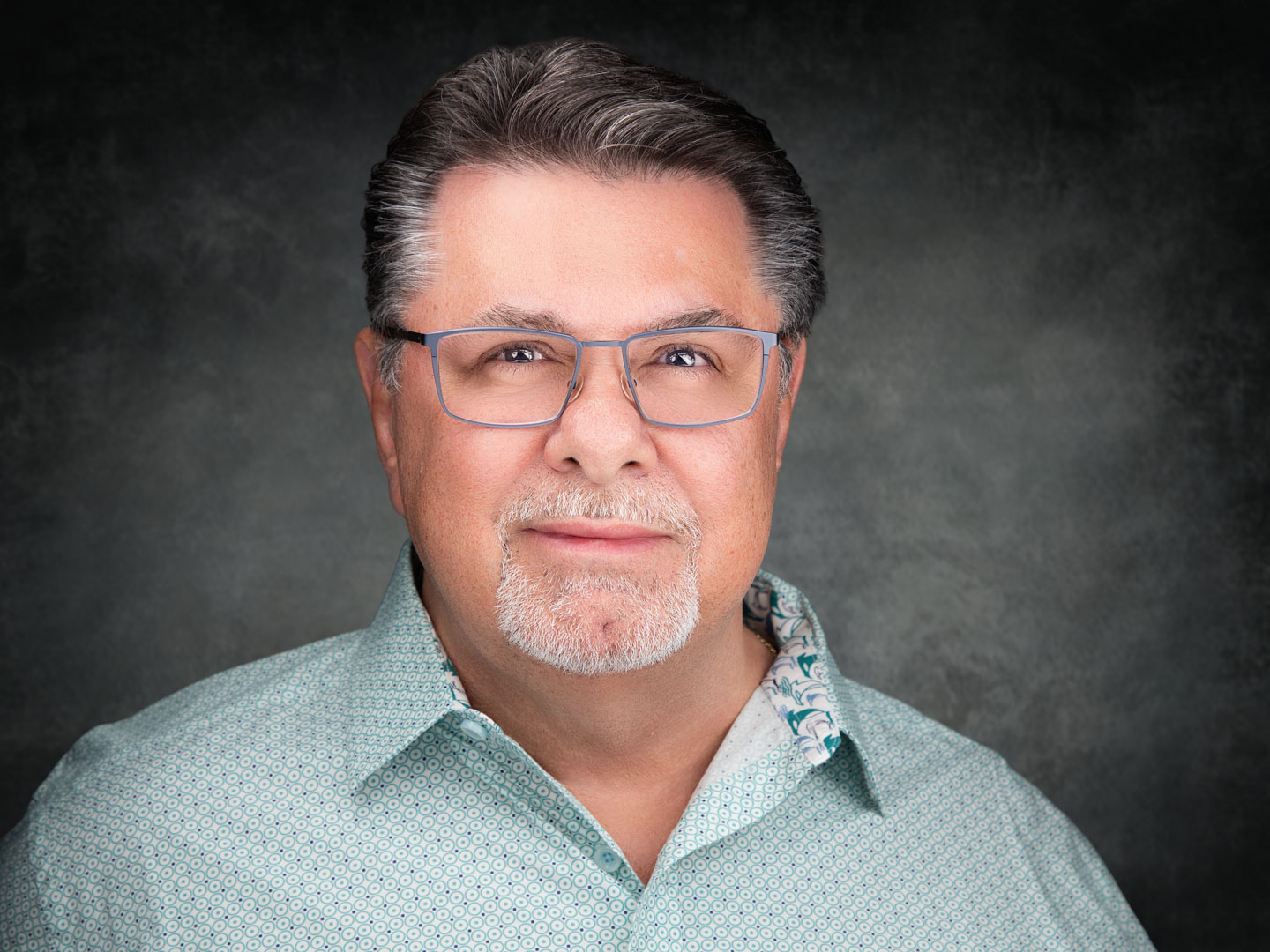 Middle-aged man with gray hair and beard wearing glasses and a light blue patterned shirt against a dark gray background.
