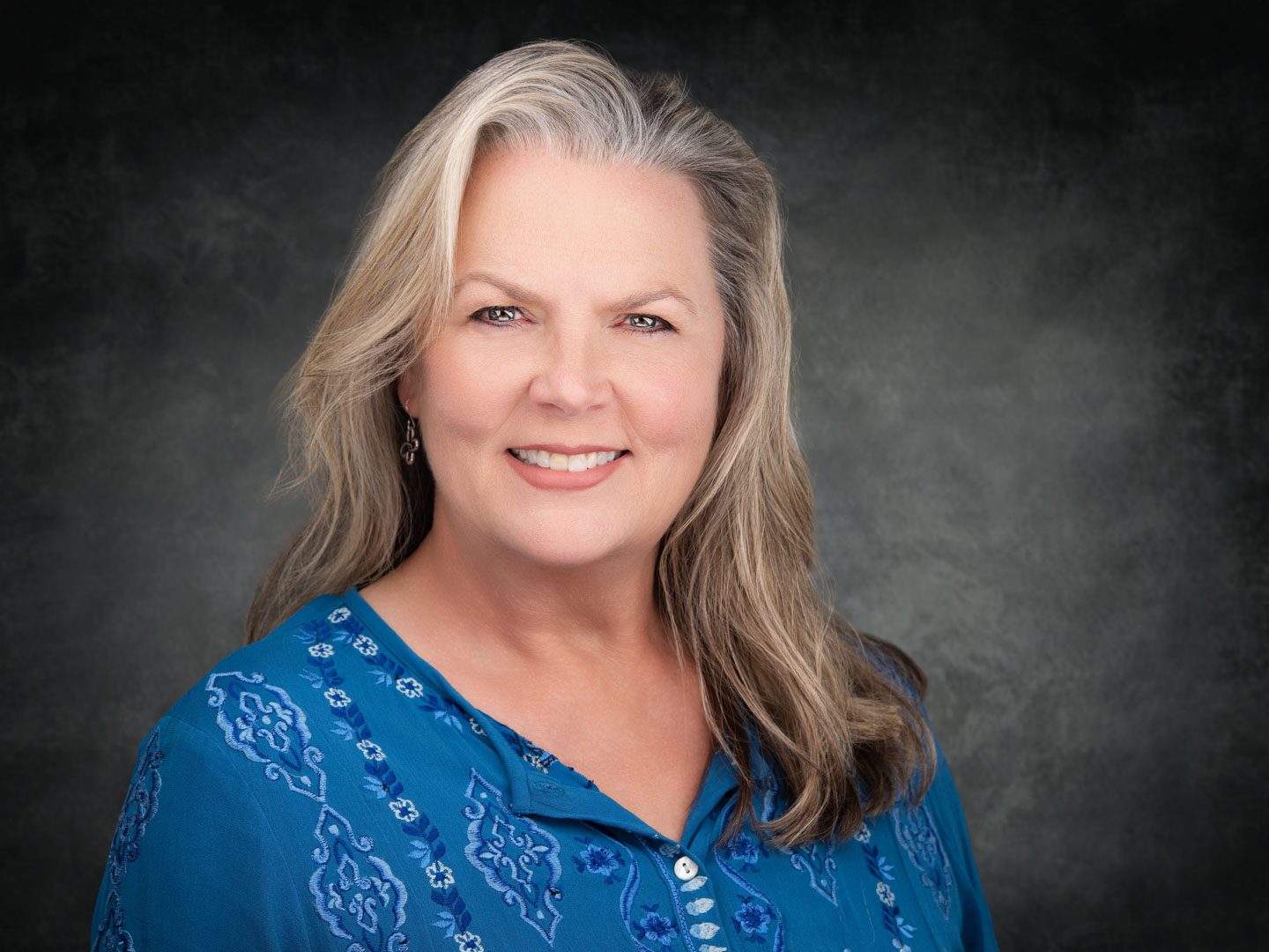 Smiling middle-aged woman with long gray hair wearing a blue patterned blouse against a dark textured background.
