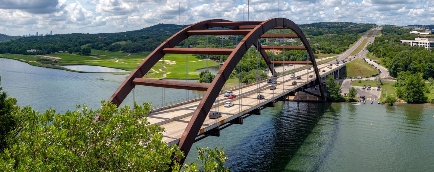 Austin 360 Bridge over a river with vehicles crossing and green landscape under a partly cloudy sky.