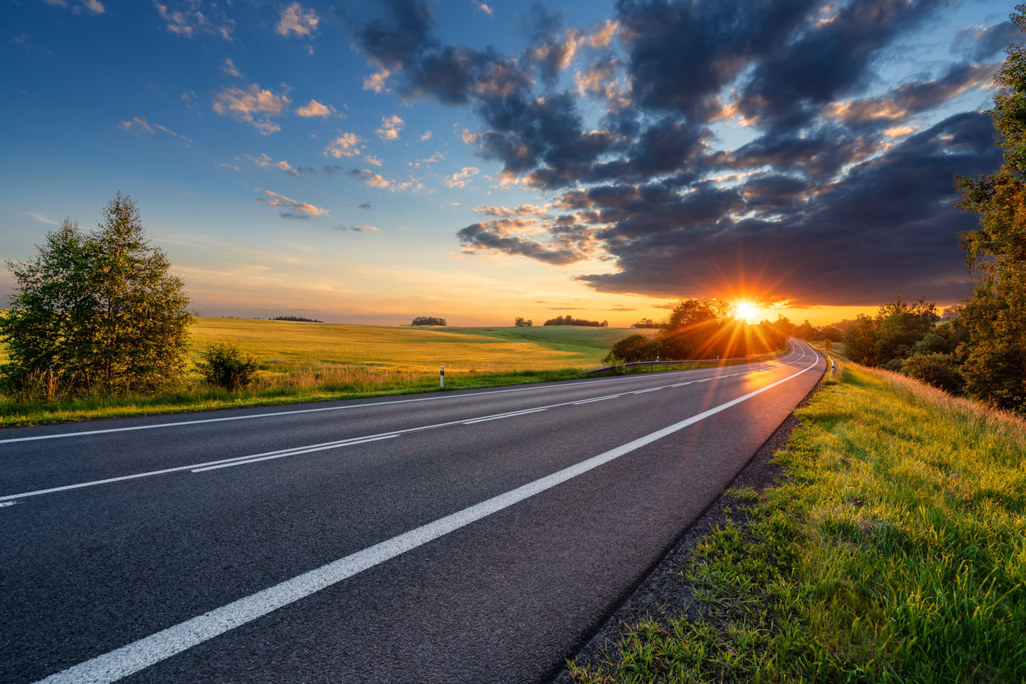 Empty asphalt road curving through a rural landscape with green fields and trees during a sunset with dramatic clouds.